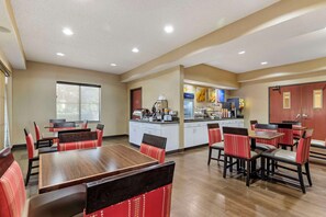 A hotel dining area with wooden tables and red upholstered chairs, a kitchen in the background, and a large window providing natural light.