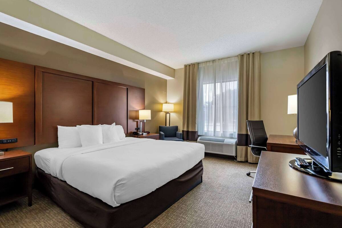 A hotel room with a large bed, a desk with a TV, a chair, a lamp, and a window with curtains.