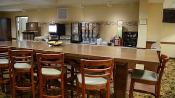 Un vestíbulo de hotel con un largo mostrador de recepción de madera, varios taburetes de bar, una máquina de café y un microondas.