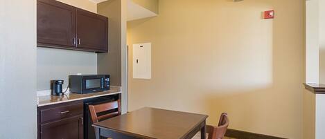 A compact kitchenette with a microwave, coffee maker, and a refrigerator. There is a small dining table with two chairs. The room has a carpeted floor and beige walls.