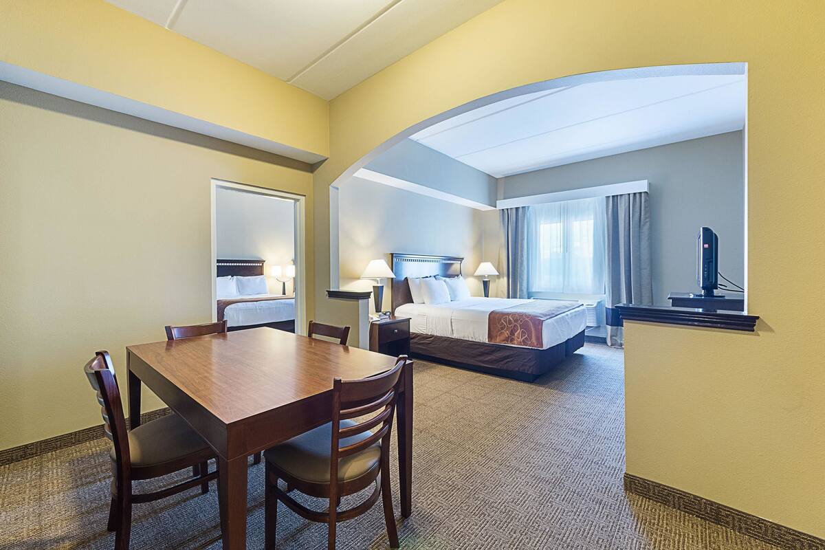 A hotel room with a wooden dining table, two chairs, a bed with a headboard, a nightstand with lamps, and a television on a stand.