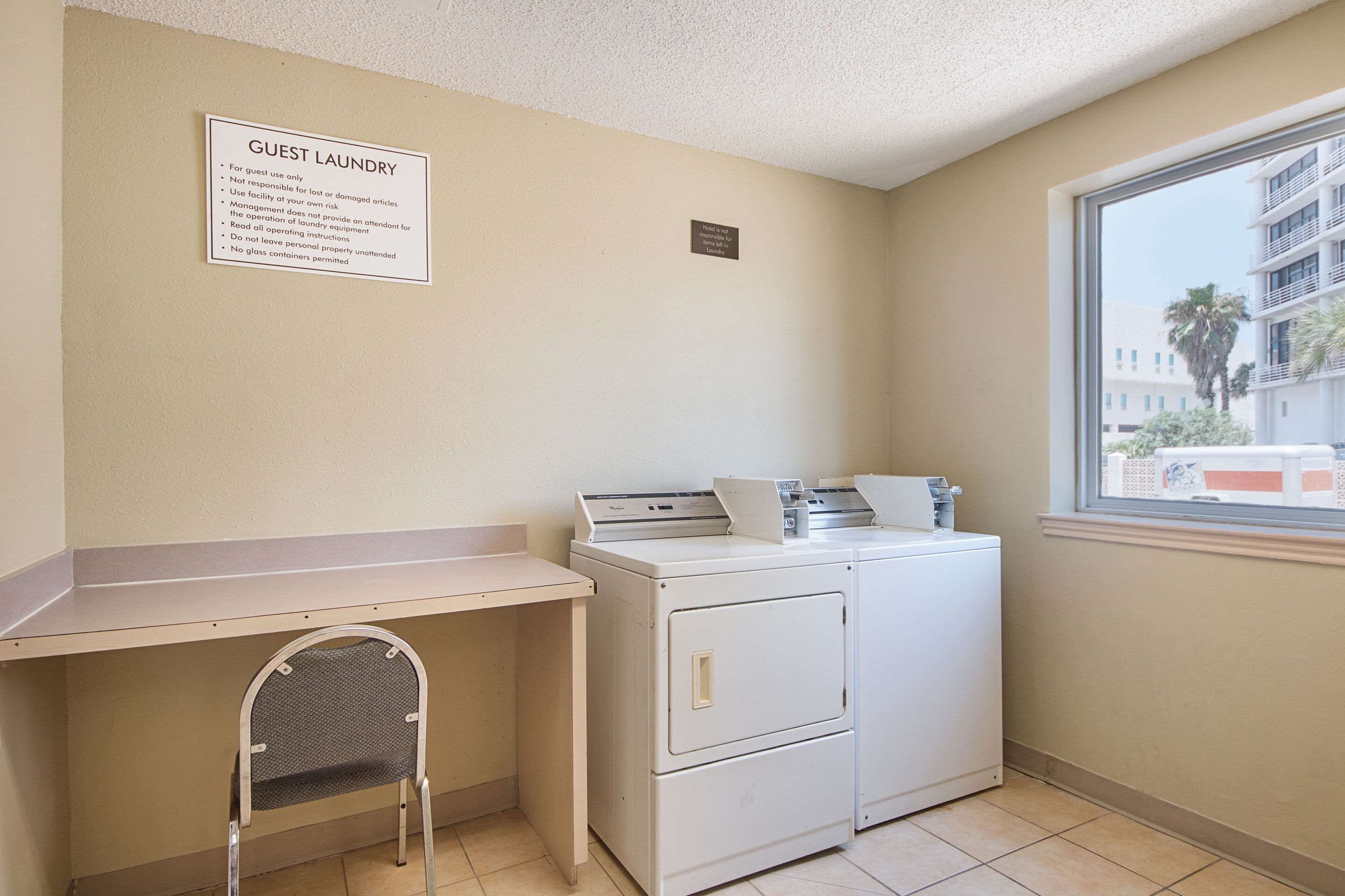 A laundry area with a sign indicating guest laundry use, a washing machine, a dryer, a chair, and a window with a view of buildings.
