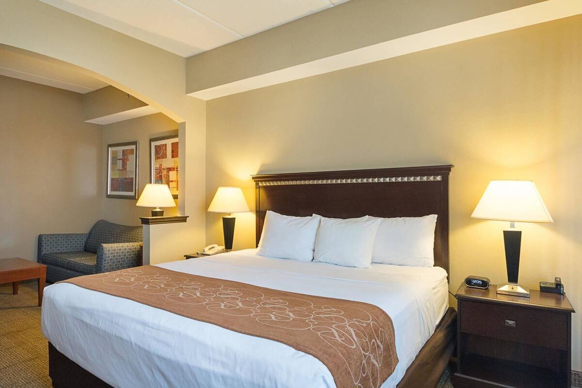 A hotel room with a large bed, two bedside lamps, a nightstand with a clock, a chair, and a small table.