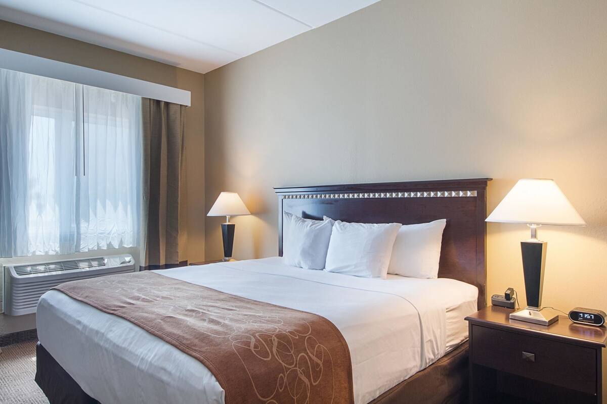 A hotel room with a large bed, two bedside lamps, a nightstand, and a window with sheer curtains.