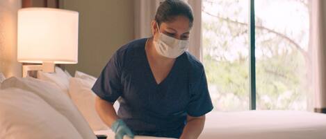 Una persona con mascarilla quirúrgica y guantes haciendo una cama en una habitación de hotel, con ropa de cama blanca y una lámpara en la mesita de noche.