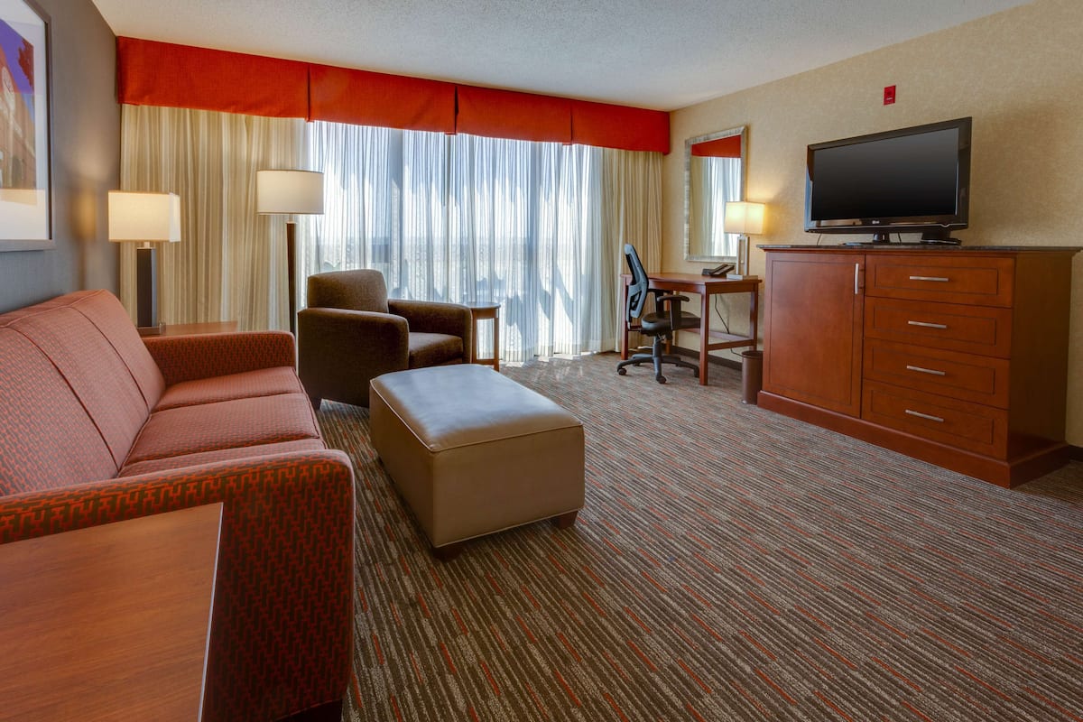 A hotel room with a sofa, ottoman, desk, chair, television, and a large window with curtains.