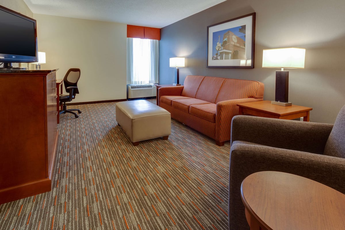 A hotel room with a television, a sofa, an ottoman, a desk with a lamp, and a framed picture on the wall.