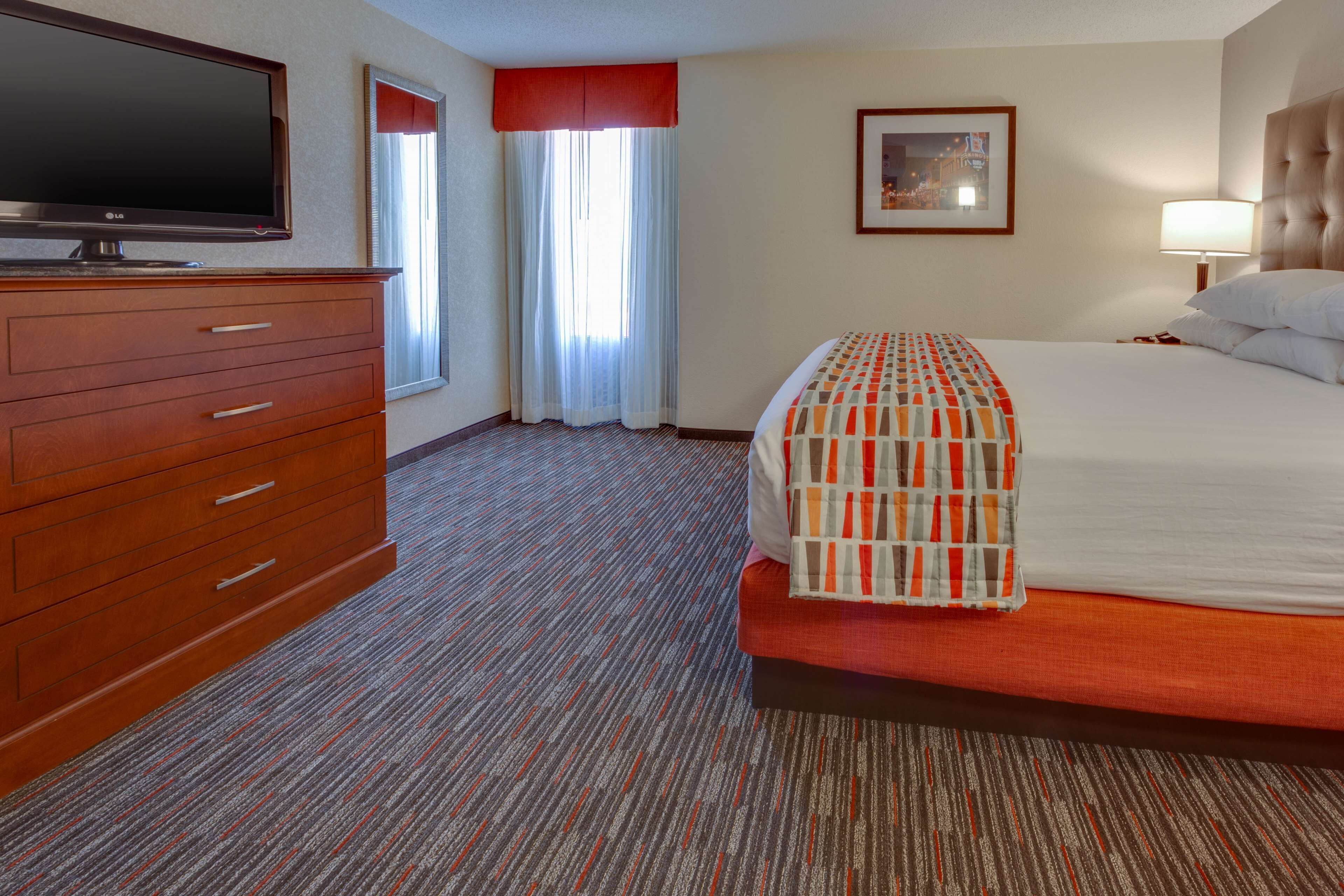 A hotel room with a bed, a TV, a dresser, a window with curtains, and a framed picture on the wall.