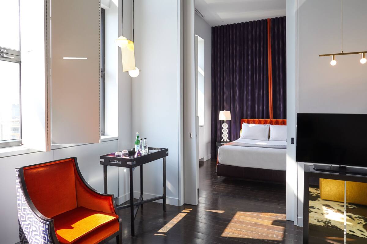 A hotel room with a bed, a TV, a desk with a lamp, a chair, and a window.