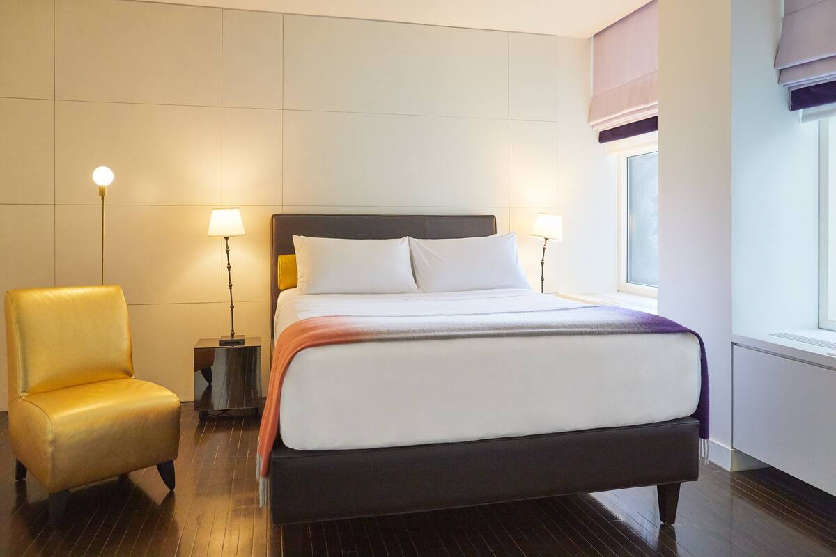 A hotel room with a bed, a chair, bedside lamps, and a window with blinds.
