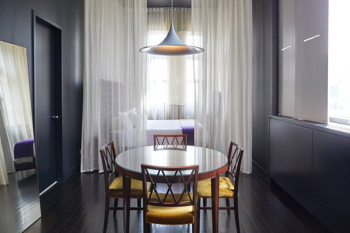 A modern dining area with a round glass table, wooden chairs with yellow cushions, a pendant light, and sheer curtains.