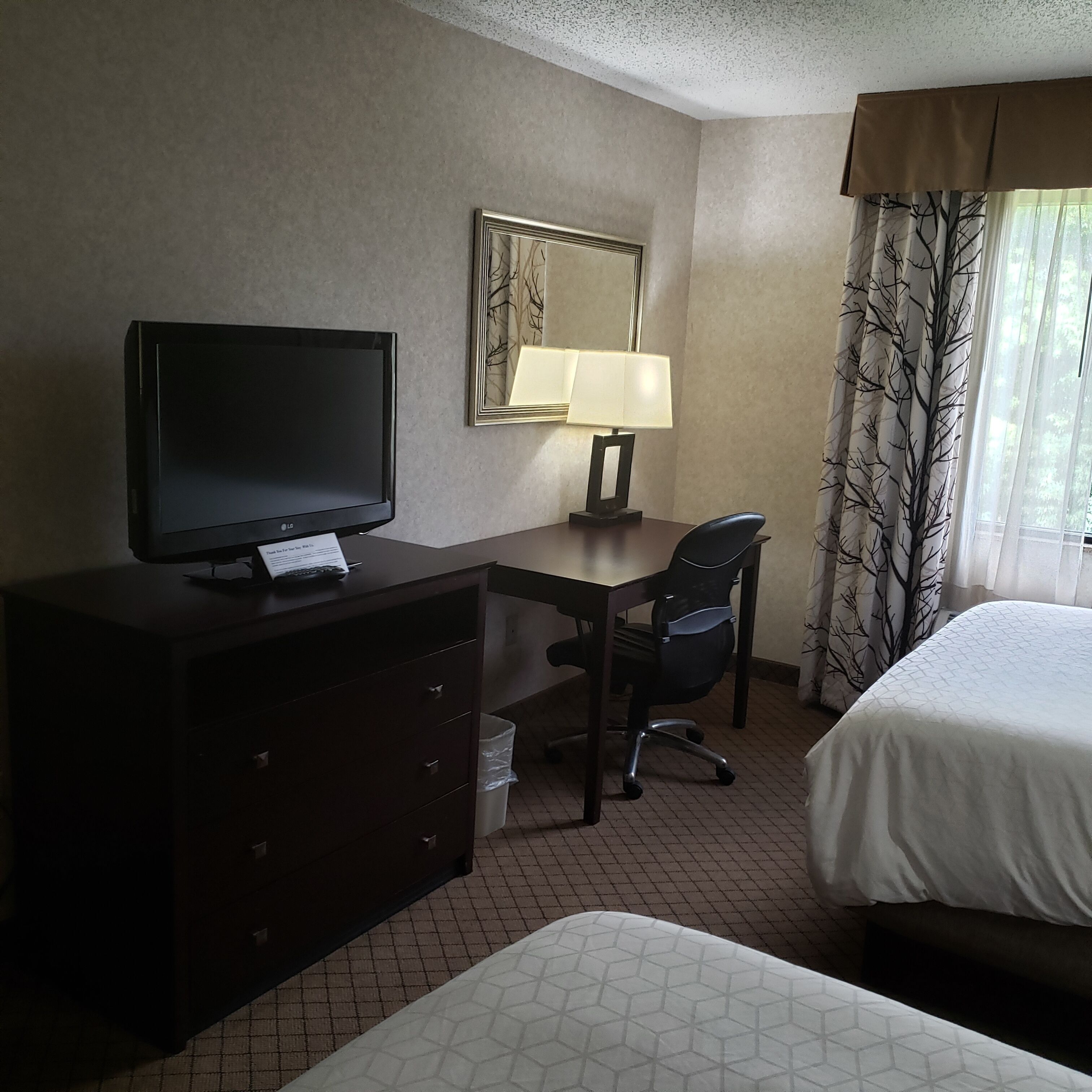 Habitación de hotel con cama, escritorio con silla, televisor, espejo y ventana con cortinas.