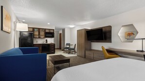 Habitación de hotel con cama, escritorio, silla, sofá, televisor y área de cocina, además de obras de arte en las paredes.