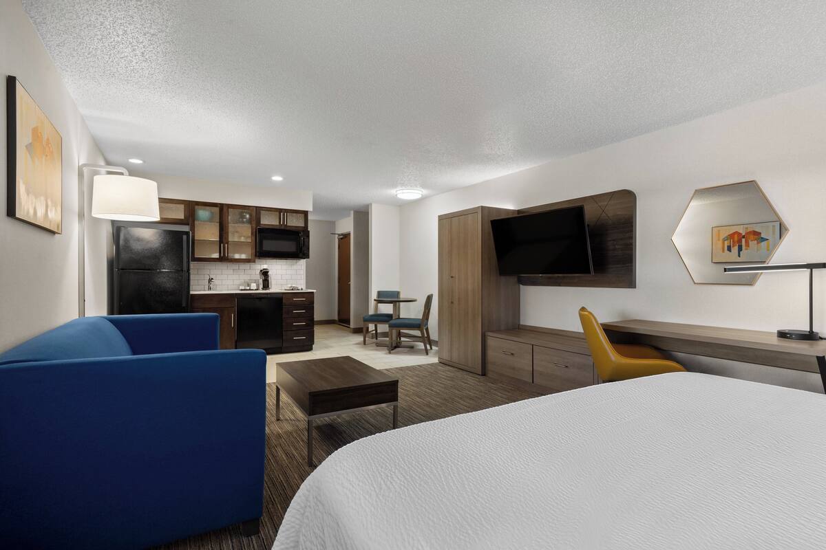 A hotel room with a bed, a desk, a chair, a sofa, a TV, a kitchen area, and artwork on the walls.