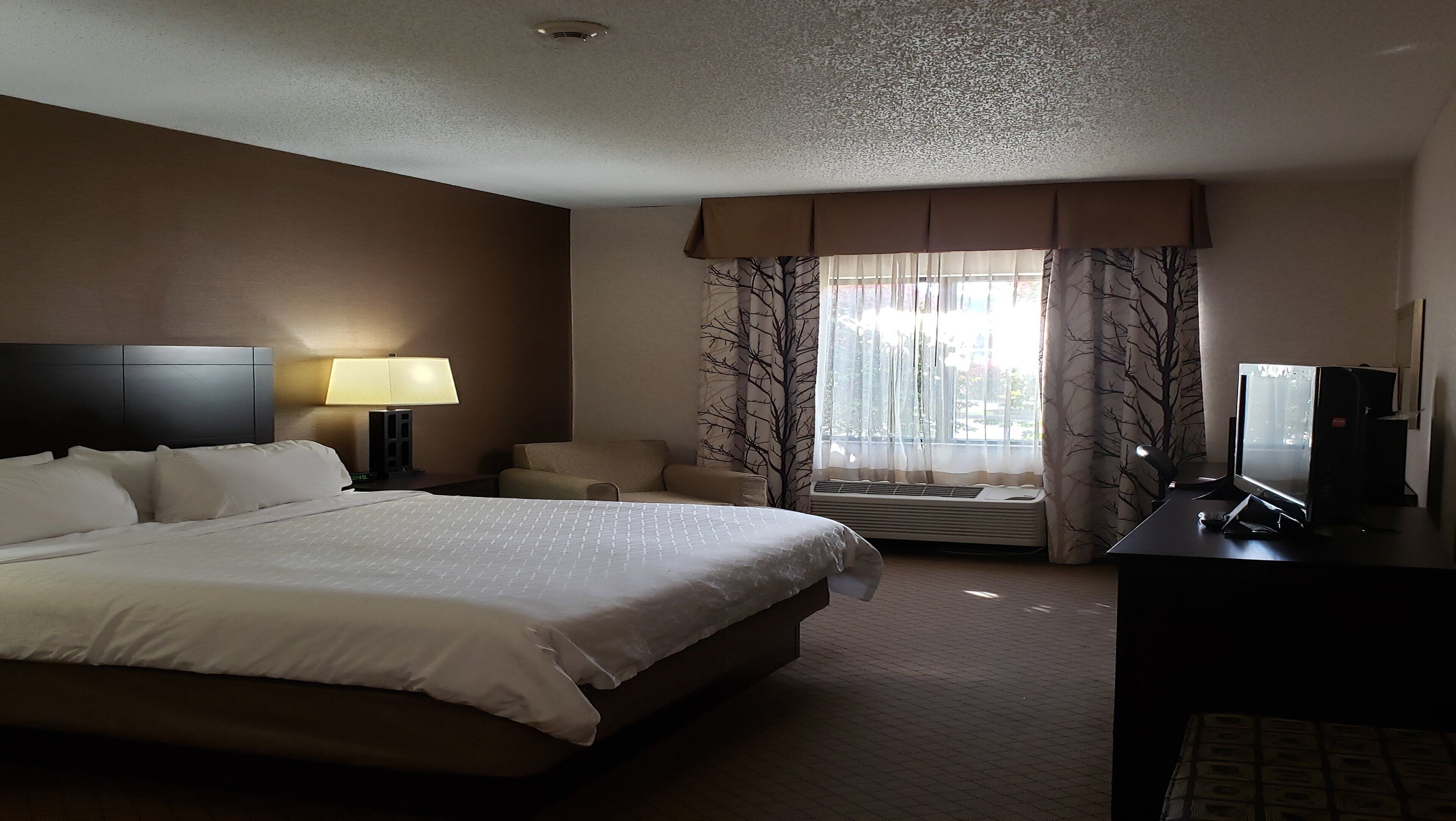 Une chambre d’hôtel avec un grand lit, un bureau avec un ordinateur, une chaise, une lampe et une fenêtre avec des rideaux.