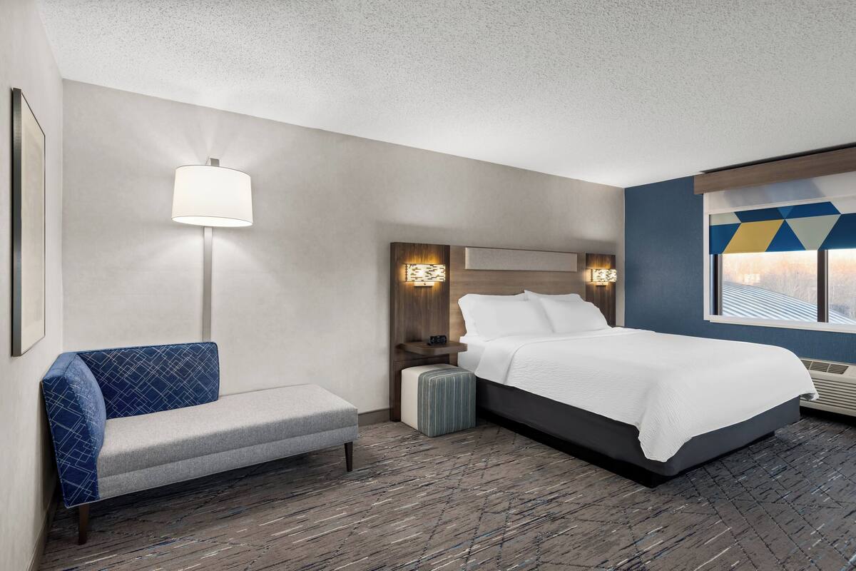 A hotel room with a bed, a bench, a nightstand, a lamp, and a window with a geometric view.
