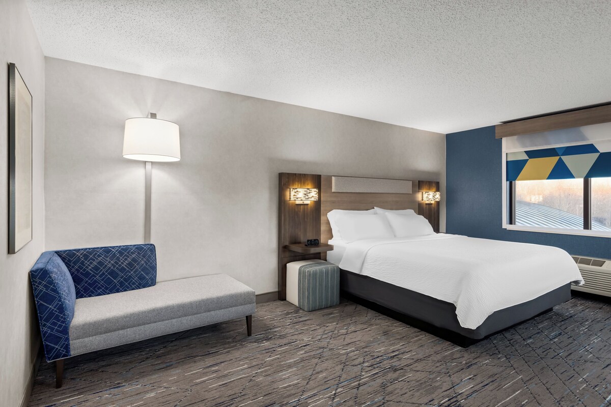 A hotel room with a bed, a bench, a wall-mounted lamp, and a window with a view.