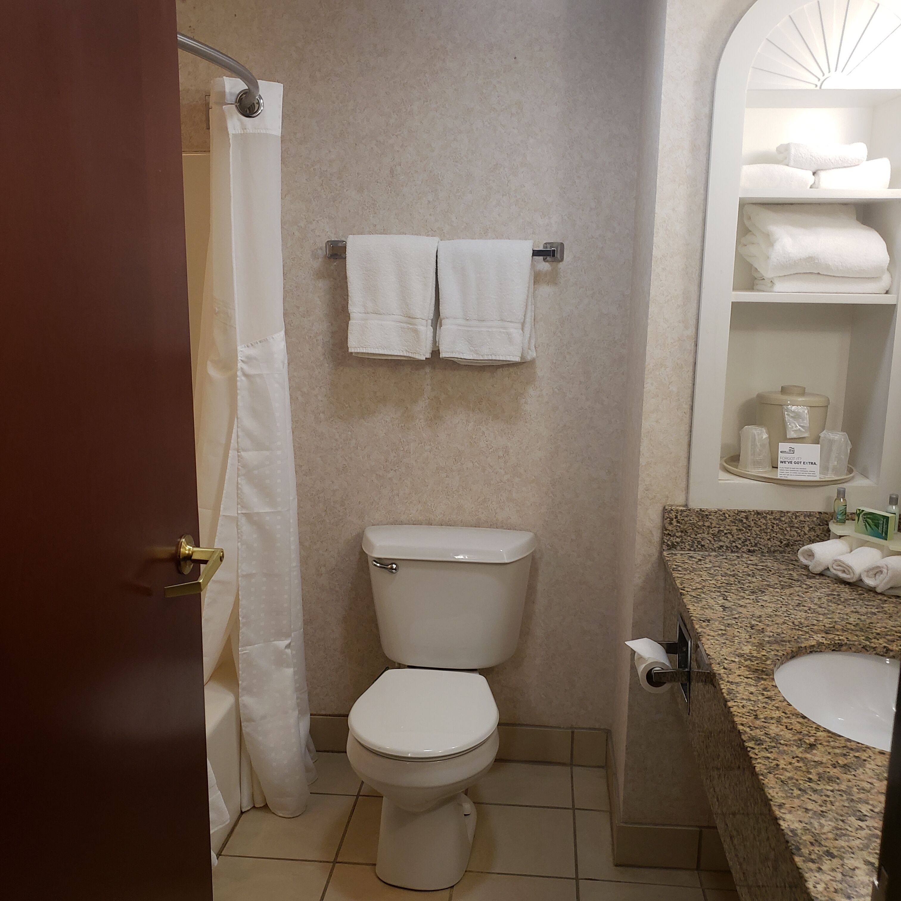 Une salle de bain comprenant des toilettes, un rideau de douche, un porte-serviettes avec deux serviettes, un lavabo avec un plan de travail en granit et une étagère contenant des articles de toilette.