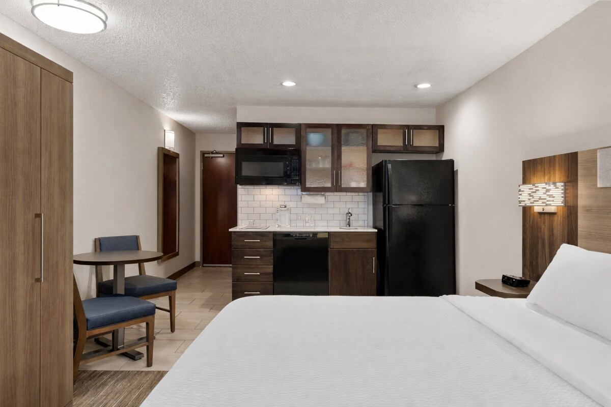 A compact hotel room with a kitchenette, a bed, a small table with chairs, and a nightstand.