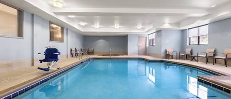 A large indoor swimming pool with a blue tiled edge, a diving board, and several chairs around the pool area.