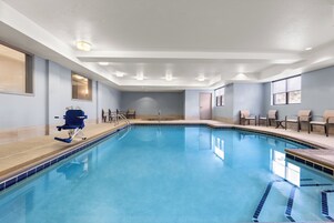 A large indoor swimming pool with a blue tiled edge, a diving board, and several chairs around the pool area.