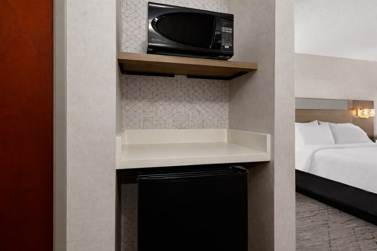 A compact kitchenette with a microwave, a small refrigerator, and a wooden shelf.