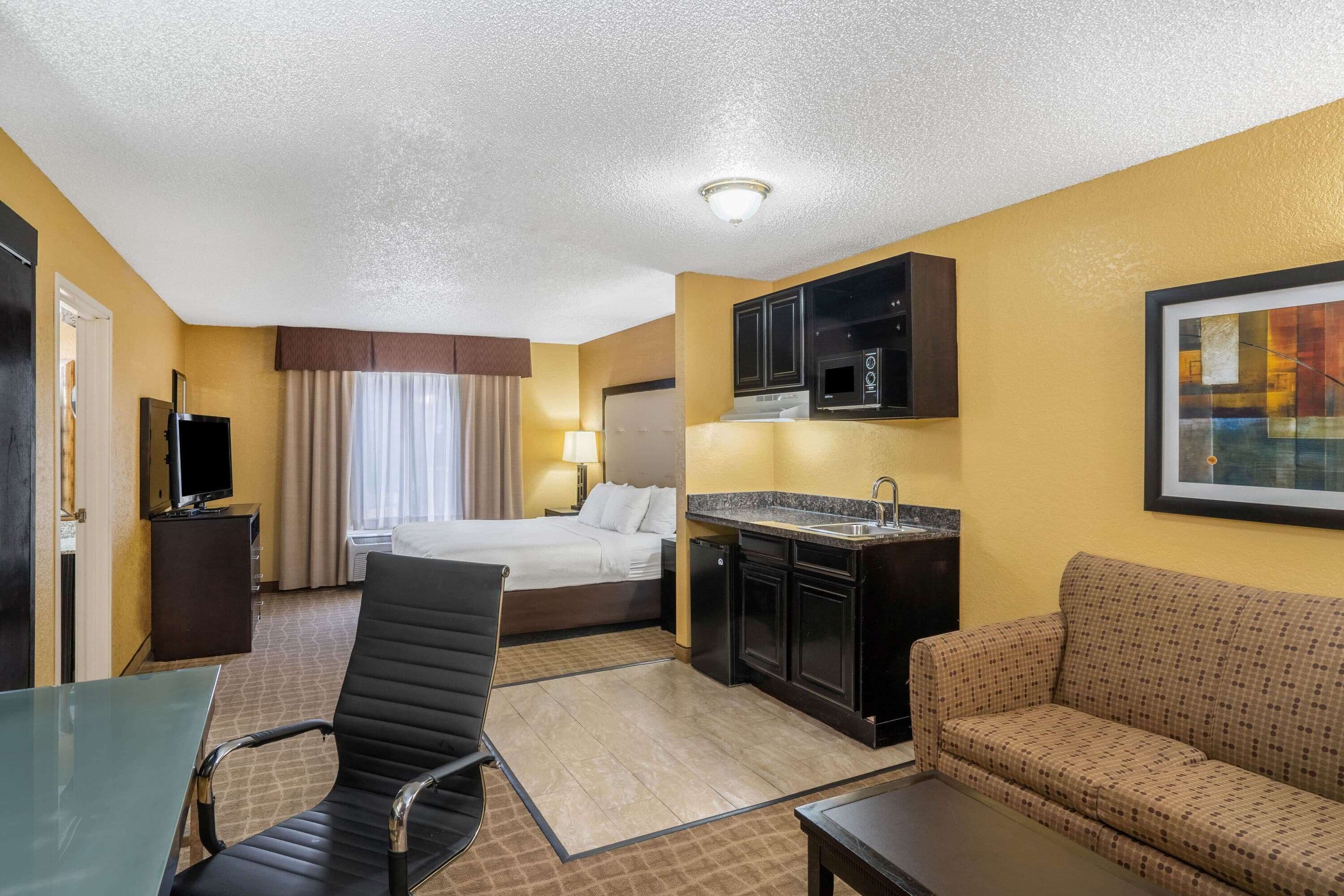 A hotel room with a bed, a desk, a chair, a sofa, a kitchenette, and a TV.