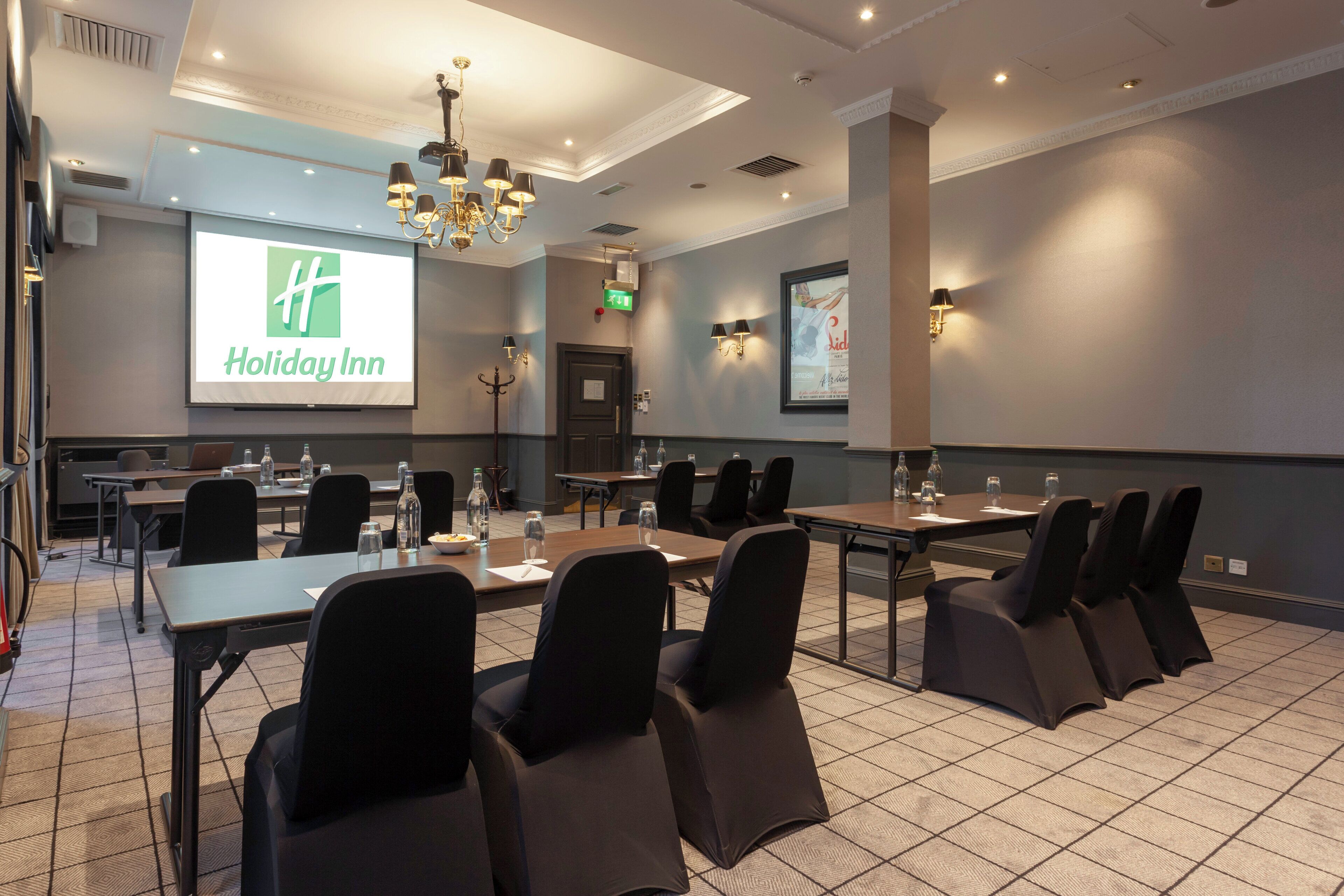 Un salón de conferencias con una pantalla grande mostrando el logotipo de Holiday Inn, varias mesas con sillas y un candelabro.