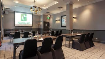 Un salón de conferencias con una pantalla grande mostrando el logotipo de Holiday Inn, varias mesas con sillas y un candelabro.