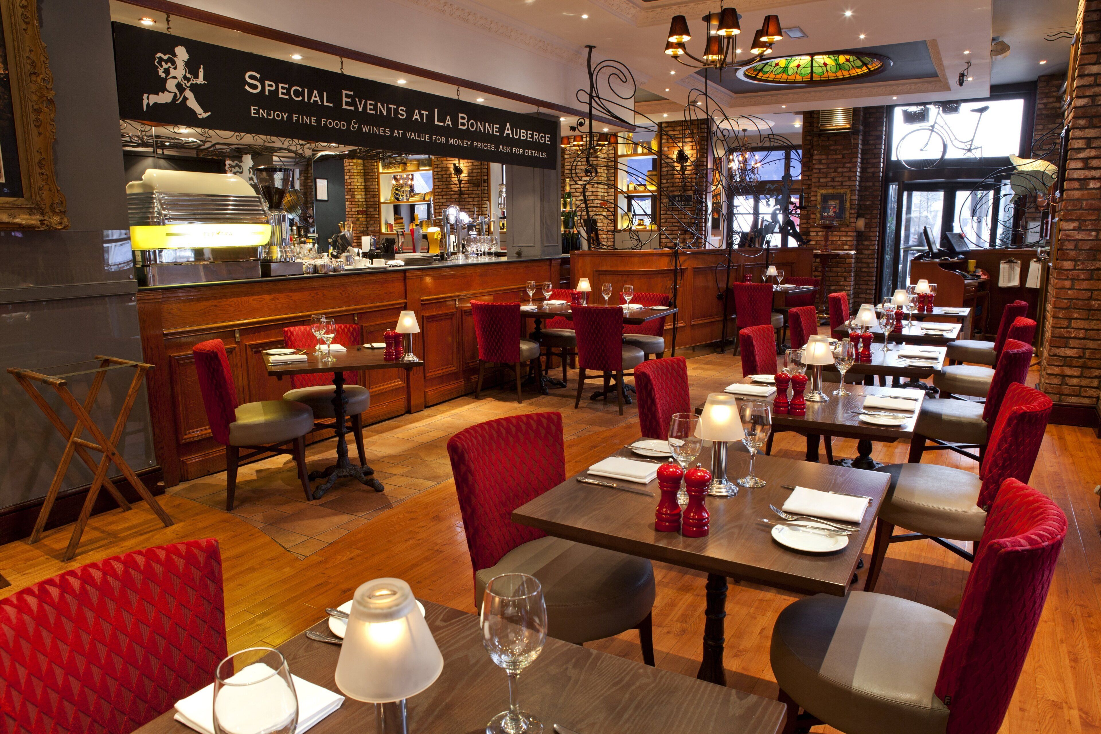 A restaurant interior with multiple tables, red upholstered chairs, and a bar area.