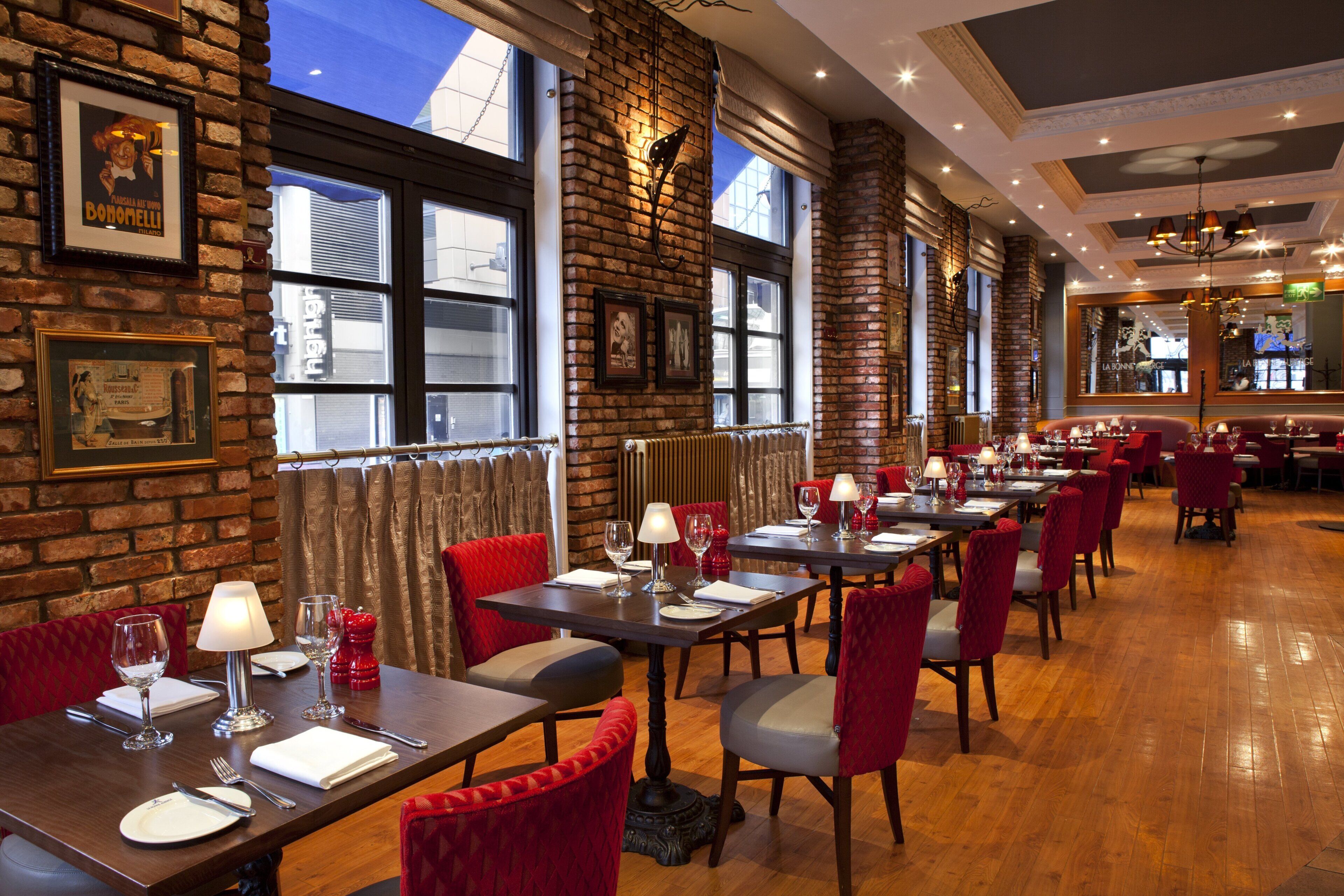 A restaurant with brick walls, large windows, and red upholstered chairs. Tables are set with wine glasses and plates.