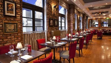 A restaurant with brick walls, large windows, and red upholstered chairs. Tables are set with wine glasses and plates.