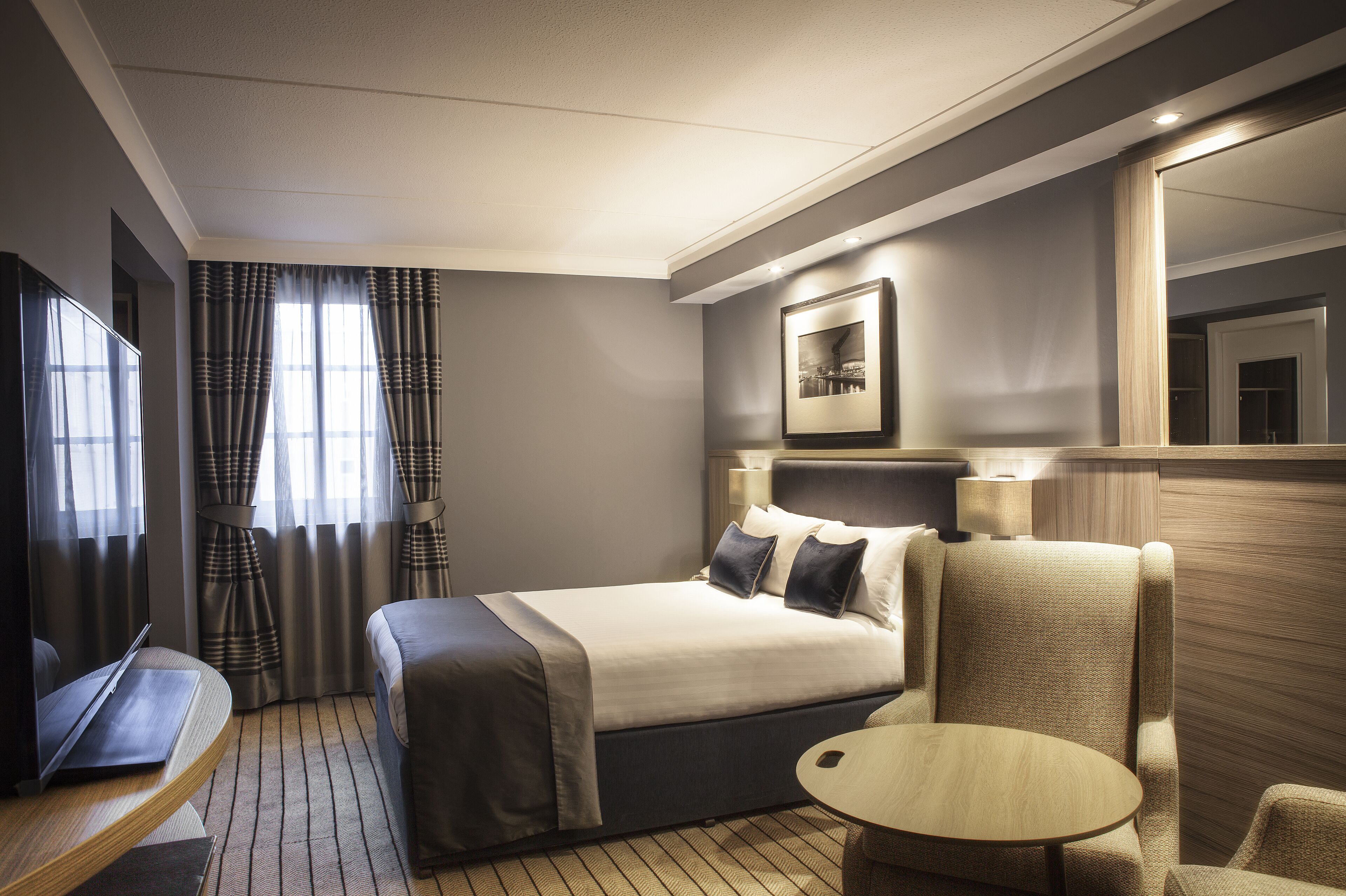 Habitación de hotel con cama, escritorio con una computadora portátil, una silla, una mesita pequeña y un espejo grande.