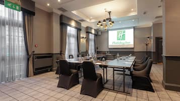 Un salón de conferencias con una amplia mesa, sillas, una pantalla de proyector que muestra el logotipo de un hotel y una araña.