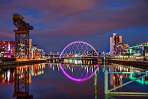 A nighttime cityscape with a prominent bridge reflecting on calm water, surrounded by illuminated buildings.