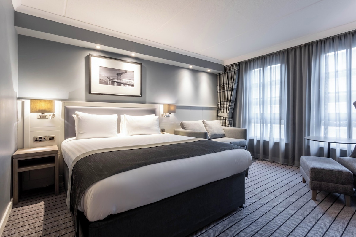 A modern hotel room with a large bed, bedside tables, a small sofa, a round table, and a large window with curtains.