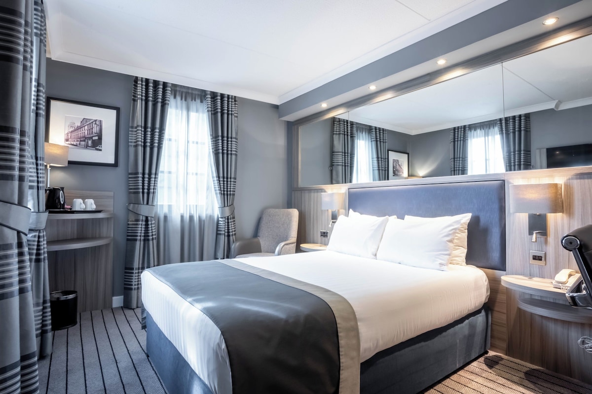 A modern hotel room with a large bed, a bedside table with a lamp, a chair, a small desk with a phone, and a large mirror above the bed.