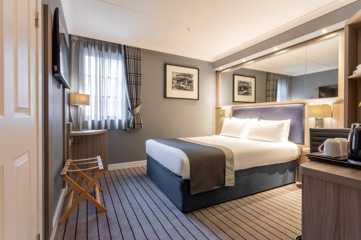 A hotel room with a bed, a desk with a lamp, a TV, a mirror, and a window with curtains.