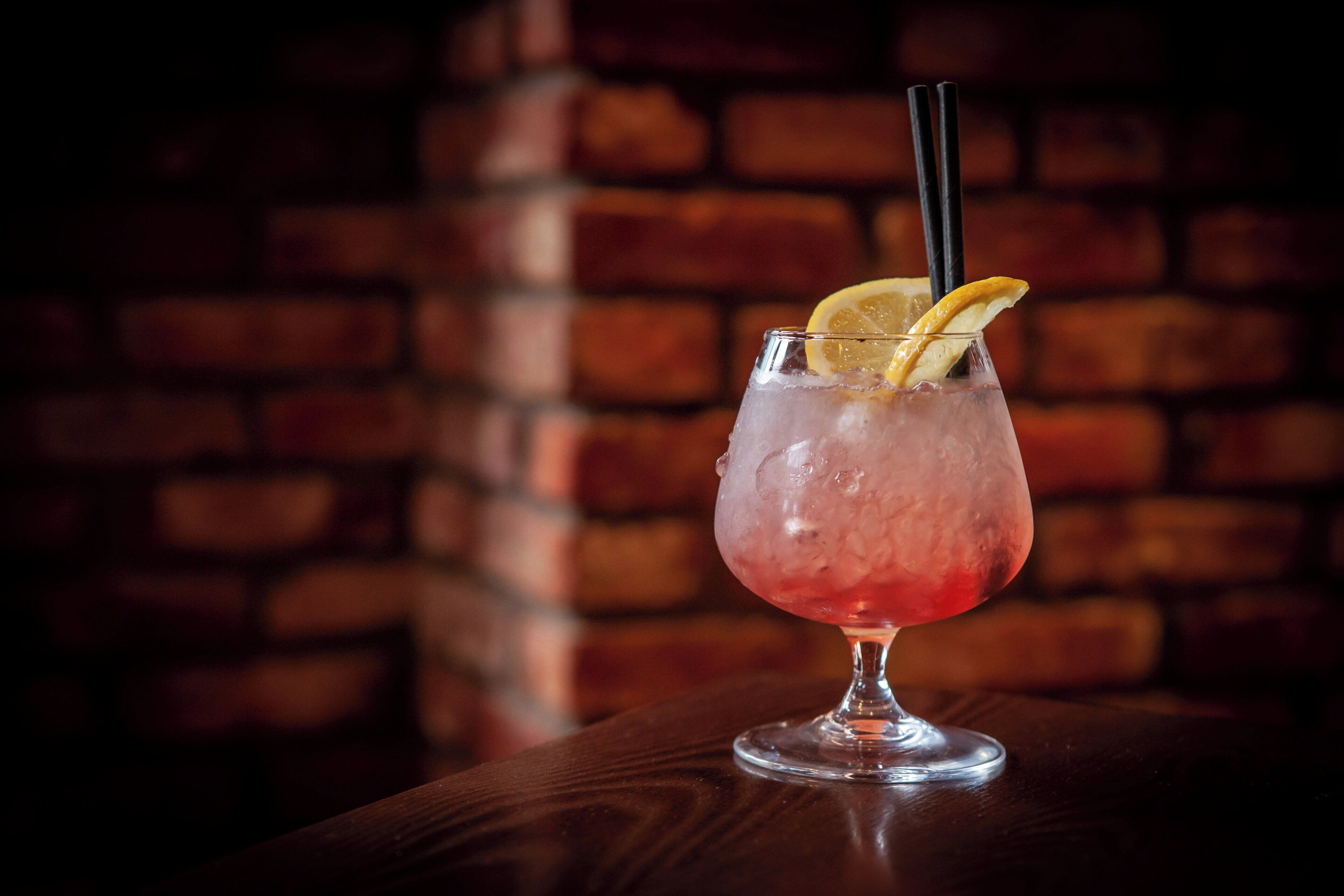 A cocktail glass with a lemon slice garnish on a wooden surface against a brick wall background.