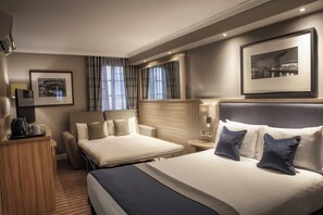 Une chambre d’hôtel avec un grand lit, un plus petit lit, un bureau et une chaise. Il y a des rideaux, un tableau encadré au mur et une lampe sur la table de chevet.