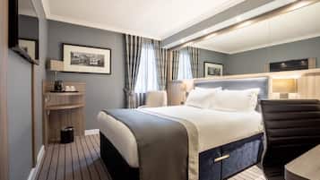 Ett modernt hotellrum med en stor säng, ett skrivbord och en stol. Det finns ett litet bord med en lampa, en tavla på väggen och en papperskorg.