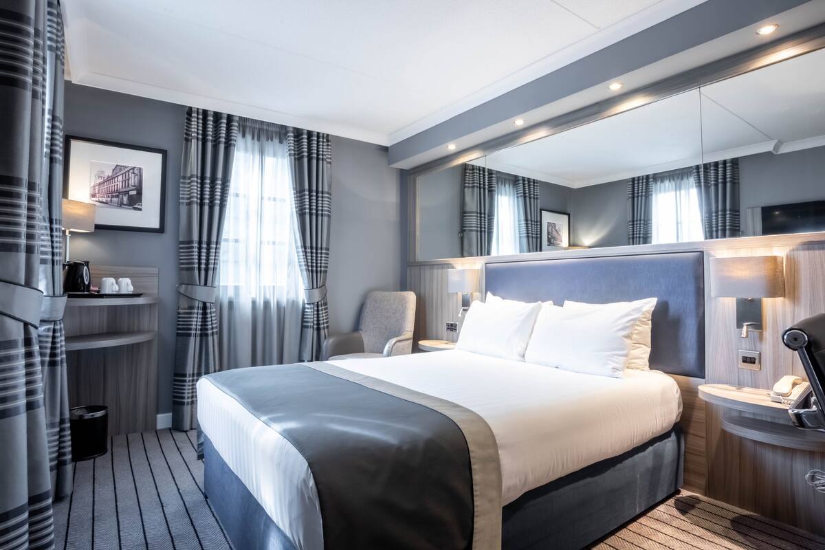 A modern hotel room with a large bed, a bedside table with a lamp, a chair, a small desk with a phone, and a large mirror above the bed.
