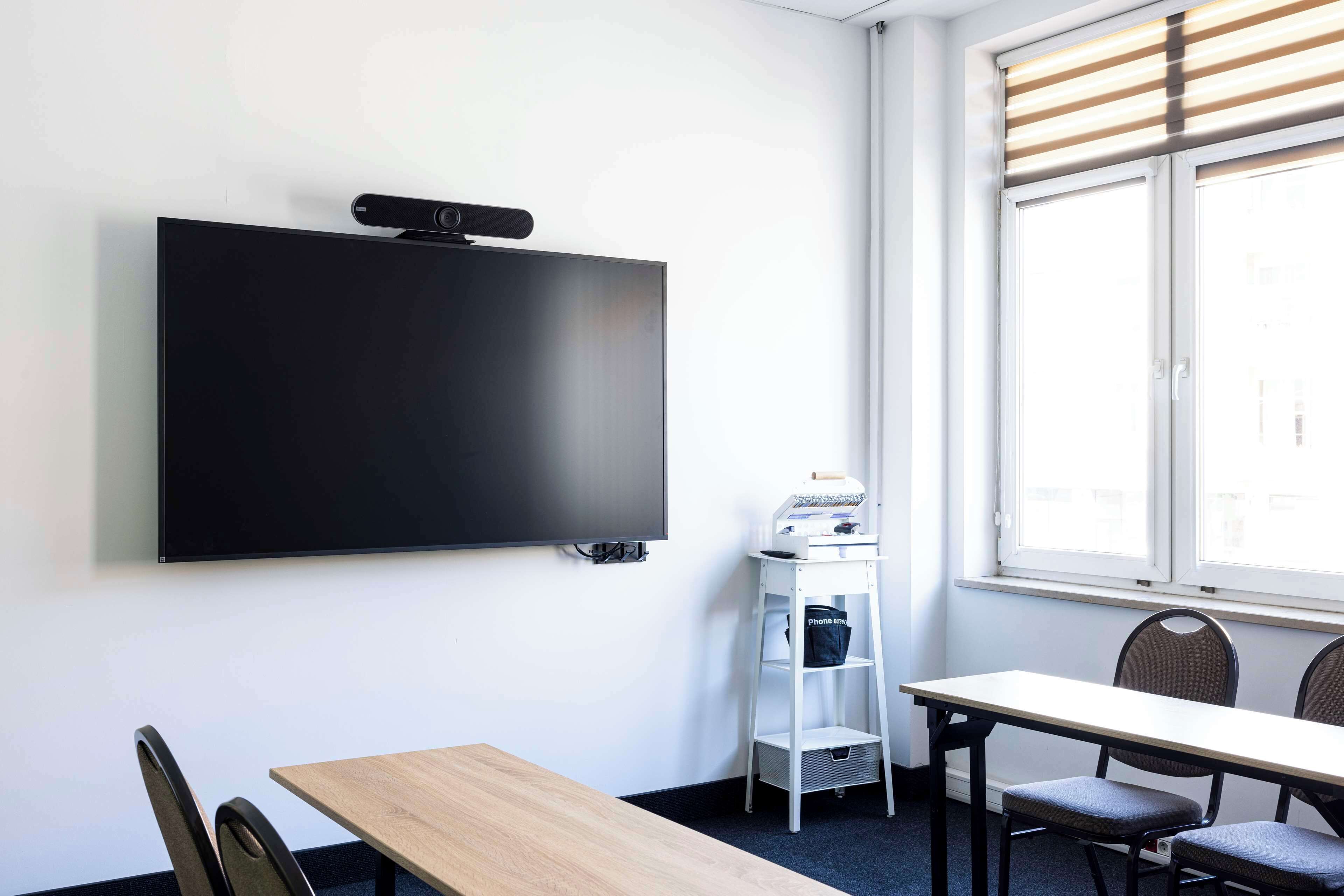 A conference room with a large flat-screen TV mounted on the wall, a whiteboard, a small white shelf with a bag, and a window with blinds.