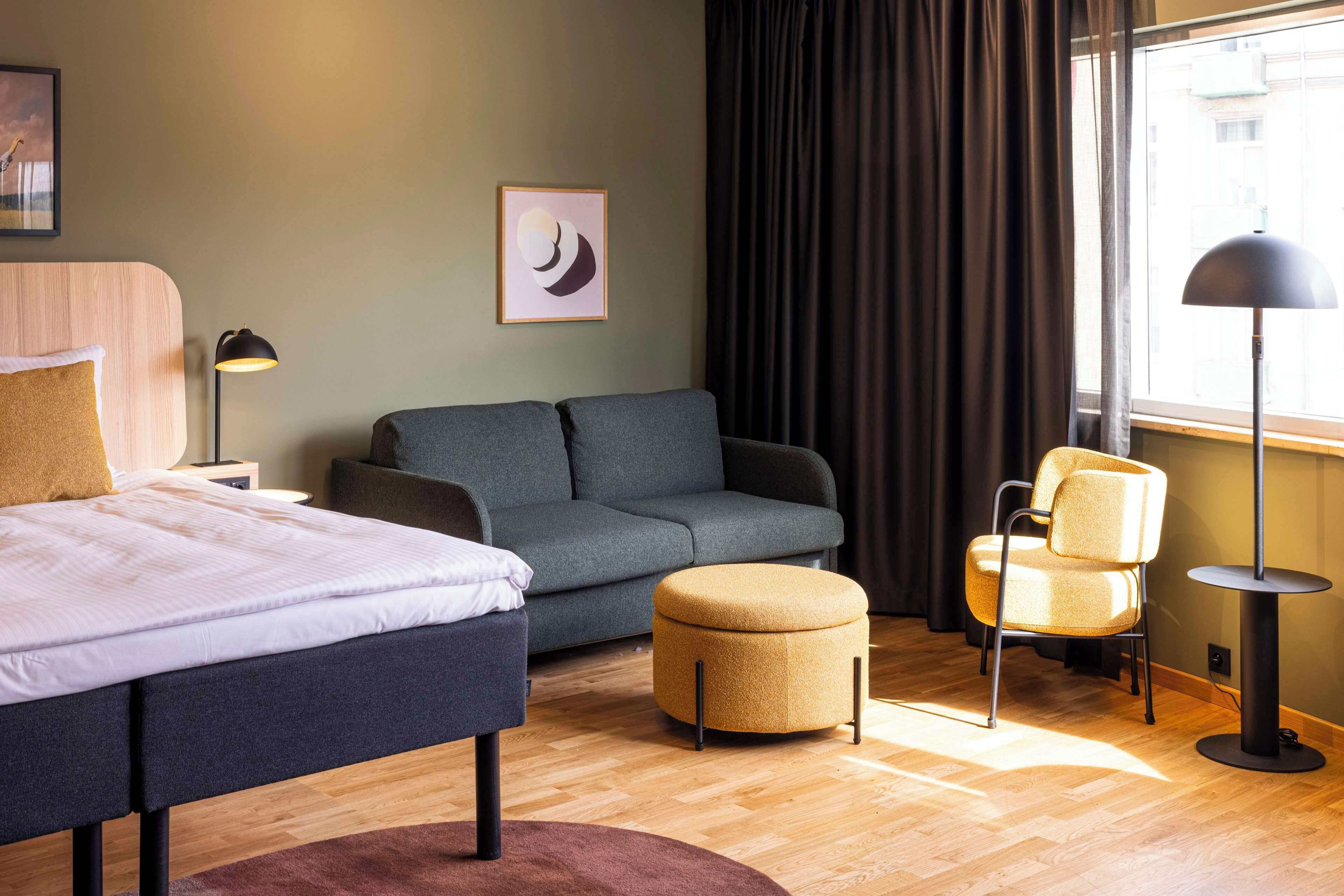 Habitación de hotel con cama, sofá, reposapiés, sillón, lámpara de pie y ventana con cortinas.