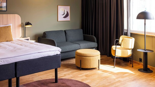A hotel room with a bed, a sofa, an ottoman, a chair, a floor lamp, and a window with curtains. - Scandic Wroclaw (Wroclaw)