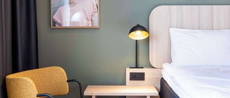 A hotel room with a bed, a desk with a lamp, a chair, and a framed picture on the wall.