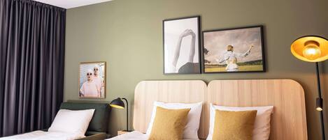 A hotel room with two beds, a large window with curtains, a wall-mounted artwork, and a bedside lamp.