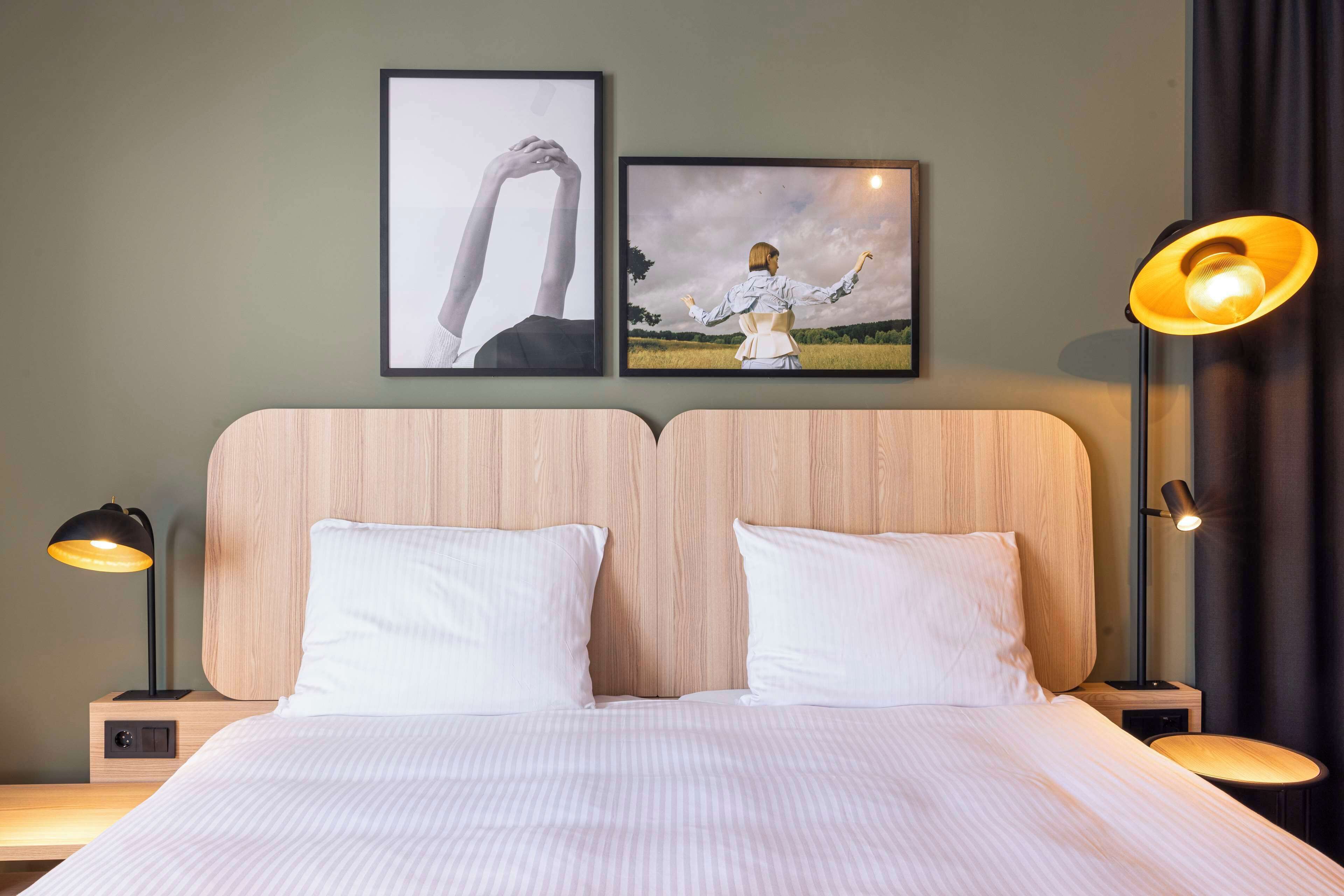 Una cama bien hecha con sábanas blancas, dos mesitas de noche con lámparas, y dos cuadros en la pared.