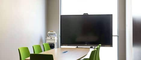 A conference room with a large flat-screen TV, green chairs, a wooden table, and a window with blinds.