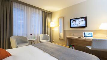 Habitación de hotel con cama, dos sillas, escritorio con una computadora portátil, televisor y ventana con cortinas.